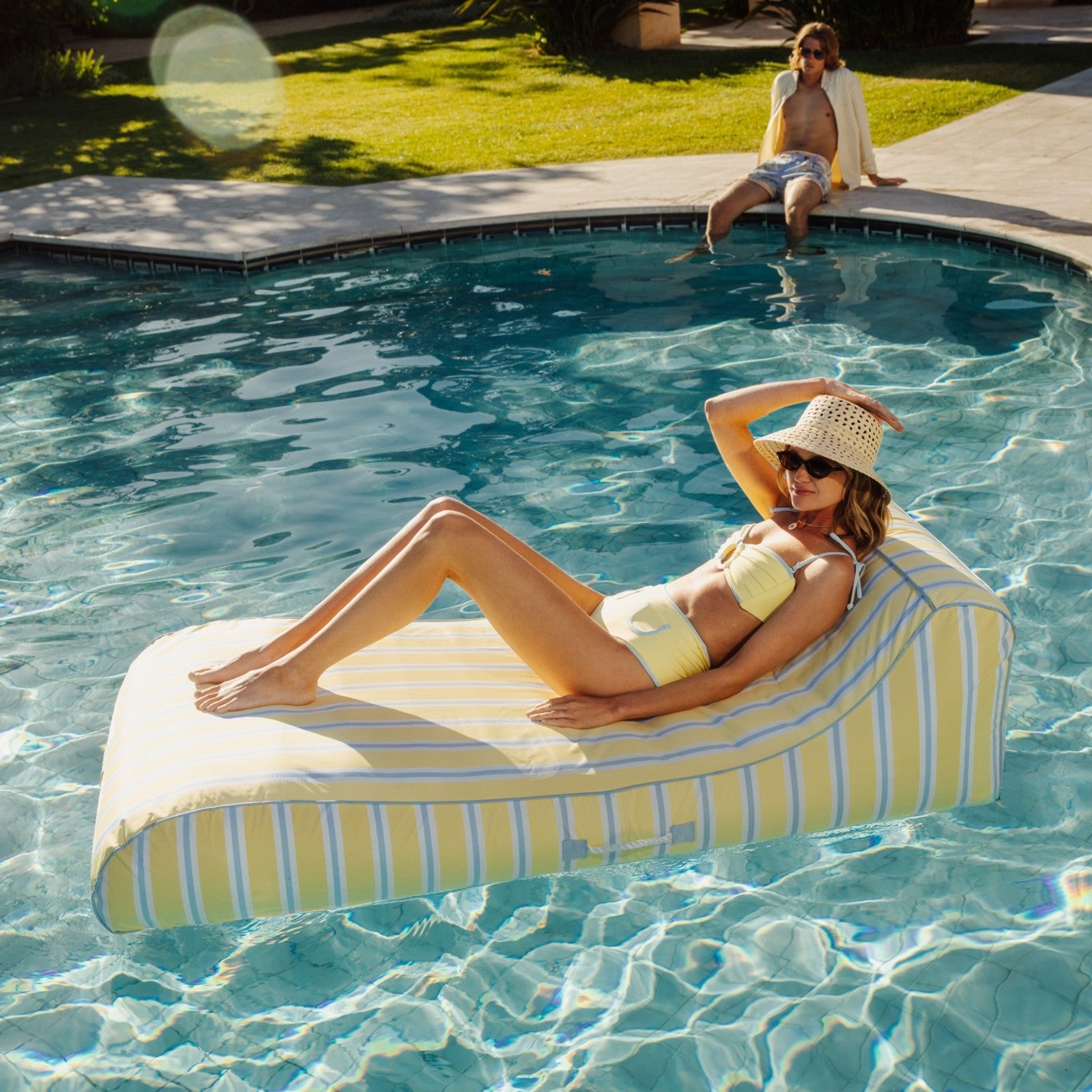 Woman lounging on a striped pool float in a swimming pool with a man sitting on the edge.