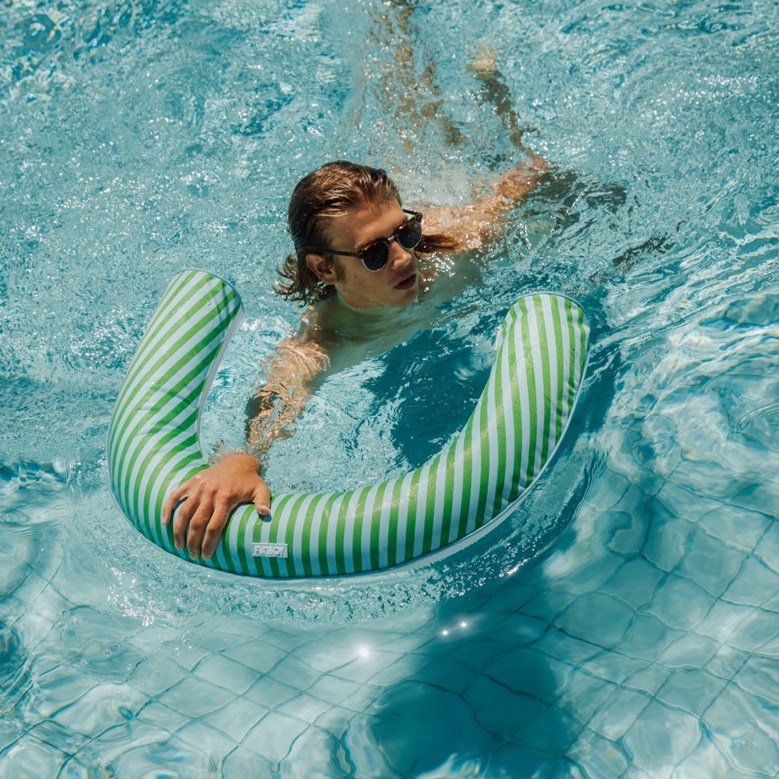 Person swimming with a green and white striped pool noodle in clear blue water.