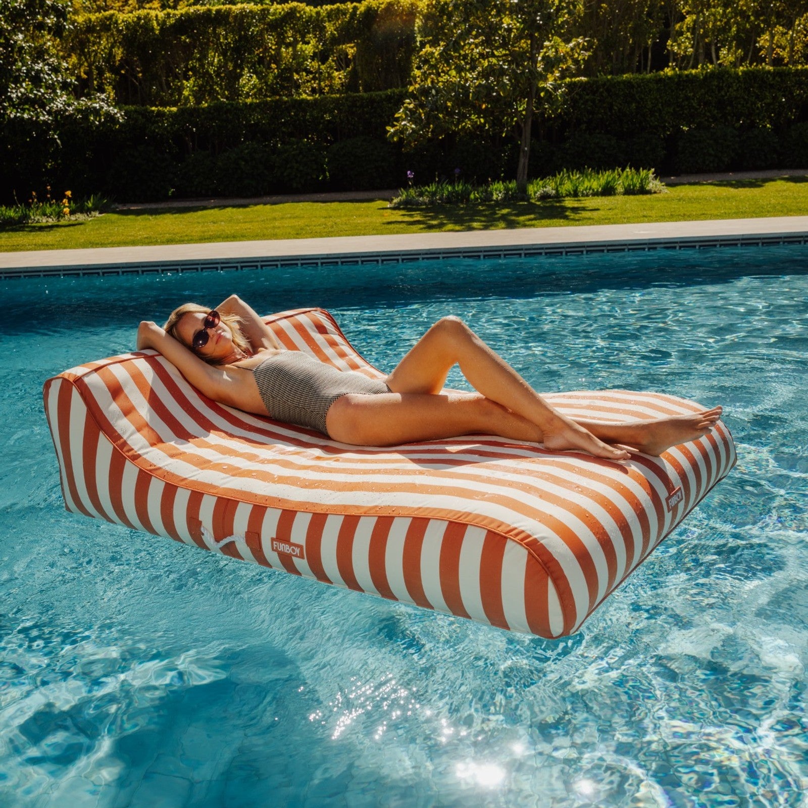 Woman relaxing on a orange striped pool float in a swimming pool with greenery in the background