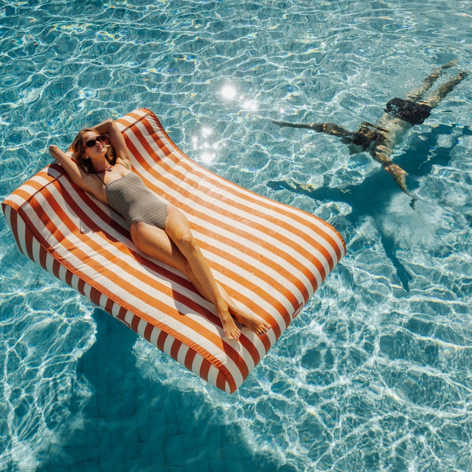 Woman lying on a striped pool float in clear blue water with a person swimming nearby.