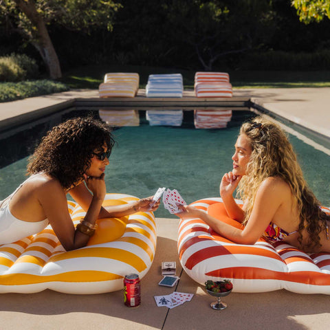 Two women playing cards on inflatable tubes by a poolside.