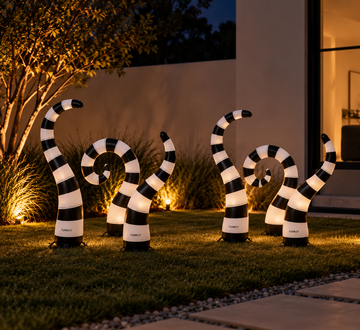 Five black and white striped garden statues shaped like stylized snakes, illuminated from within, arranged on a lawn in front of a modern house at night.