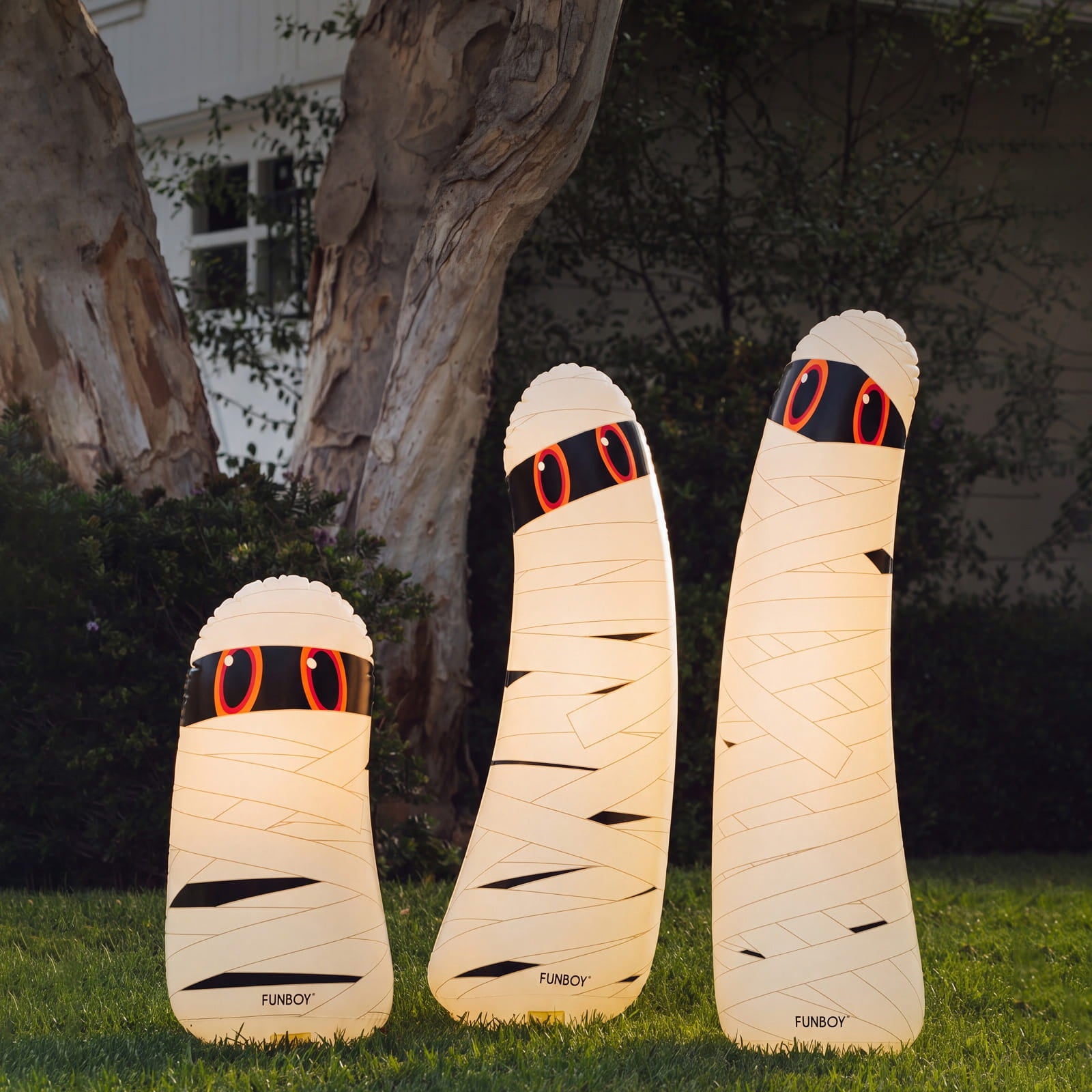 Three mummy-themed outdoor floor lamps shaped like wrapped bandages, standing on a grassy lawn in front of trees at night. The lamps glow warm white with black bandage details and red circular eye accents.