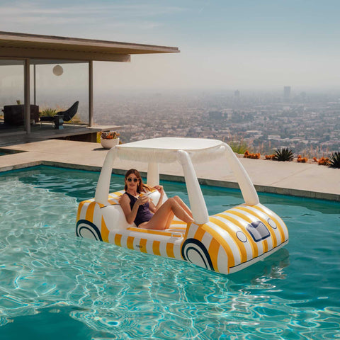 Woman lounging on a striped yellow golf cart pool float in a pool with a cityscape view.