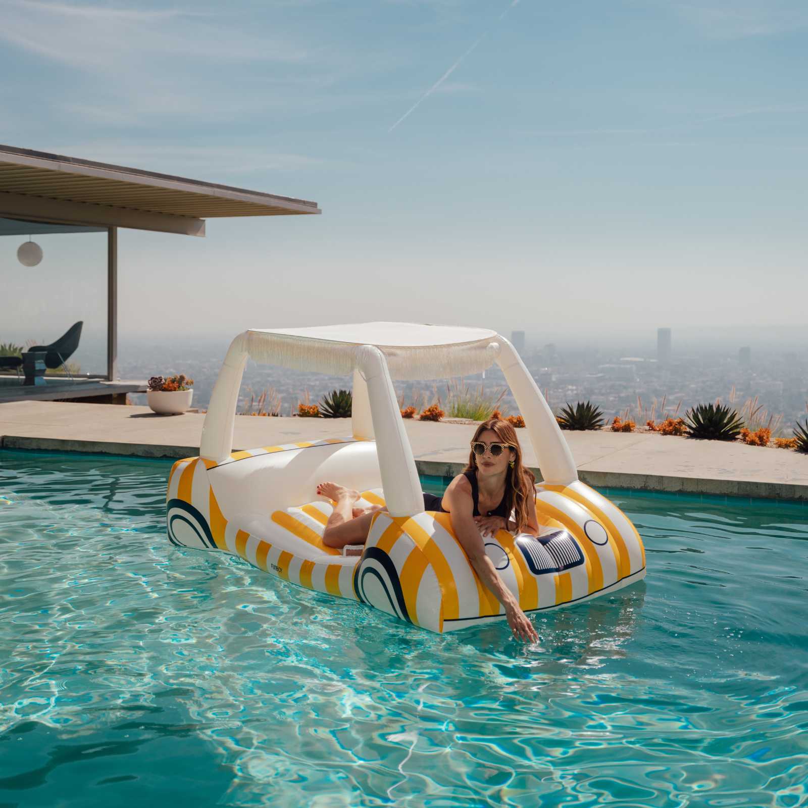 Woman lounging on a striped pool float with canopy in a pool, surrounded by a scenic view.