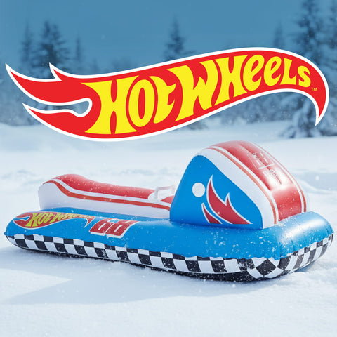 A Hot Wheels themed inflatable snowmobile slide toy resting on a snowy ground with pine trees in the background. The product features a blue base with a black and white checkered pattern, red and white accents, and a blue headrest with a red flame design.