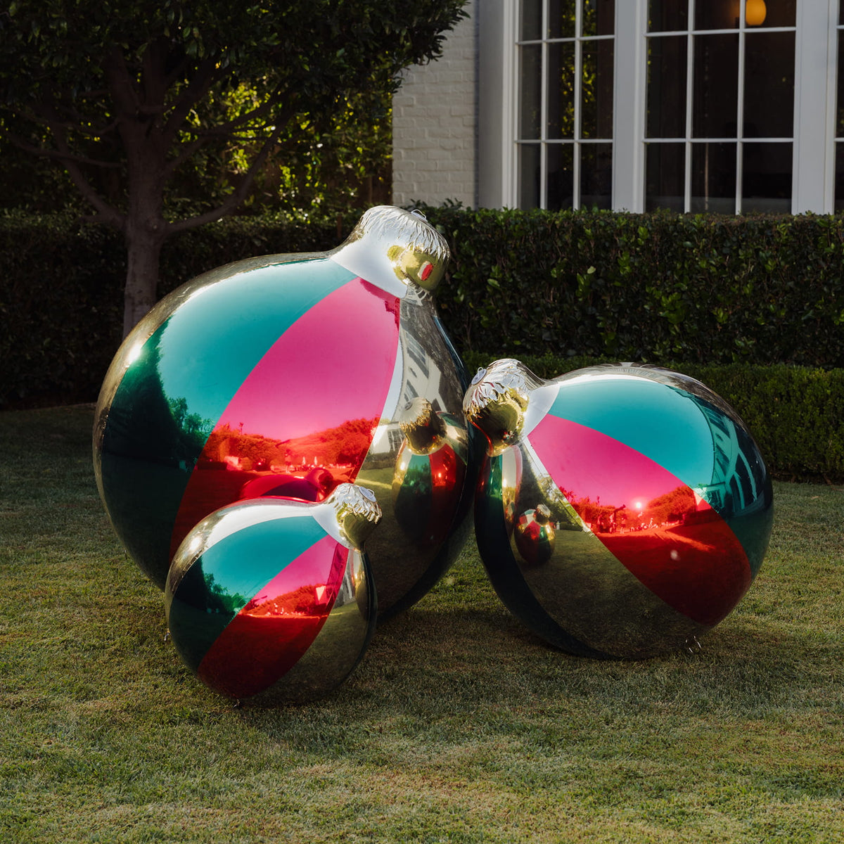 Three large reflective holiday ornaments in metallic silver with diagonal pink, teal, and red panels, displayed on a grassy lawn in front of a house and hedge.