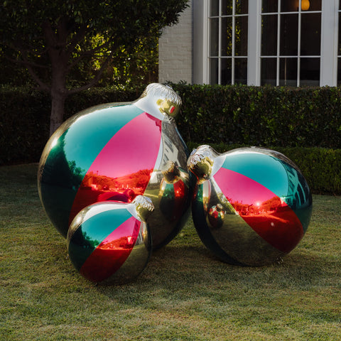 Three large reflective holiday ornaments in metallic silver with diagonal pink, teal, and red panels, displayed on a grassy lawn in front of a house and hedge.