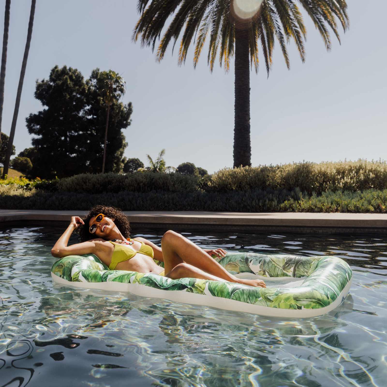 Woman lounging on a tropical-patterned inflatable raft in a pool with palm trees in the background