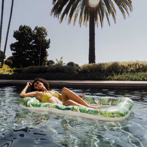 Woman lounging on a tropical-patterned inflatable raft in a pool with palm trees in the background