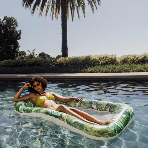 Woman lounging on a leaf-patterned inflatable pool float in a pool with palm trees in the background.
