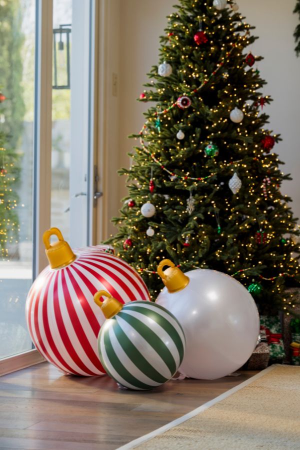 Three oversized Christmas ornaments in red and white stripes, green and white stripes, and solid white, displayed on a wooden floor in front of a decorated Christmas tree.