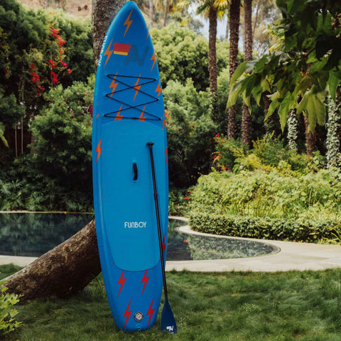 Blue paddleboard with red lightning bolt designs on a grassy area with greenery in the background