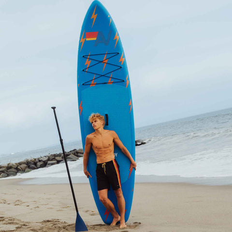 Man holding a blue paddleboard with red and yellow designs on a beach. FUNBOY x Aviator Nation
