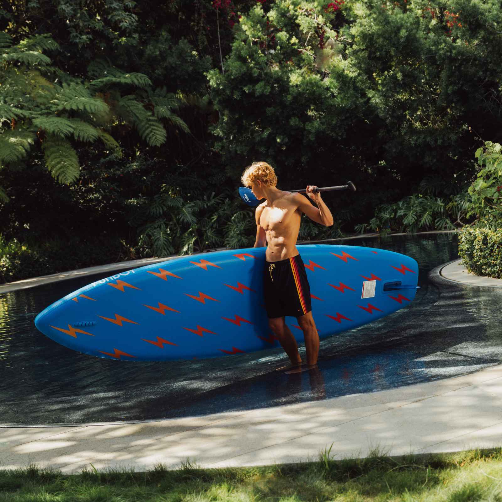 Man holding a blue paddleboard with orange patterns near a pool. FUNBOY x Aviator Nation