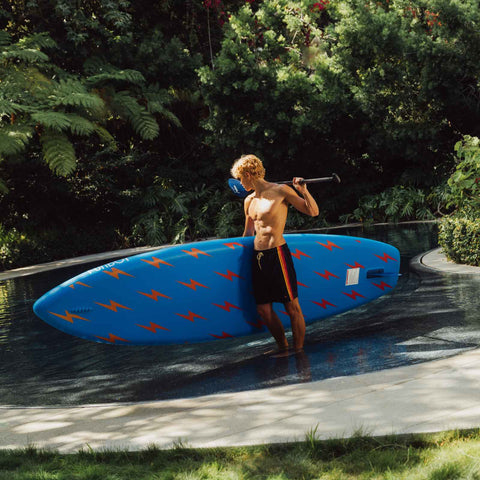 Man holding a blue paddleboard with orange patterns near a pool. FUNBOY x Aviator Nation