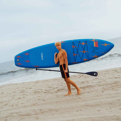 Man carrying a blue paddleboard with red graphics on a beach. FUNBOY x Aviator Nation