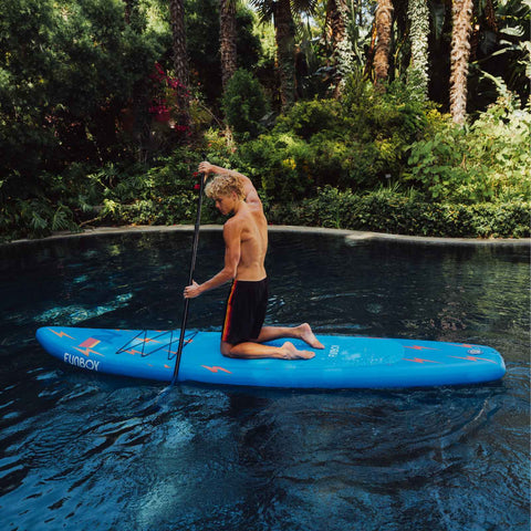 Man on a blue paddleboard in a pool surrounded by greenery. FUNBOY x Aviator Nation