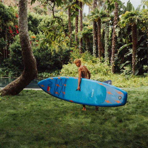 Person holding a blue paddleboard in a lush green garden. FUNBOY x Aviator Nation