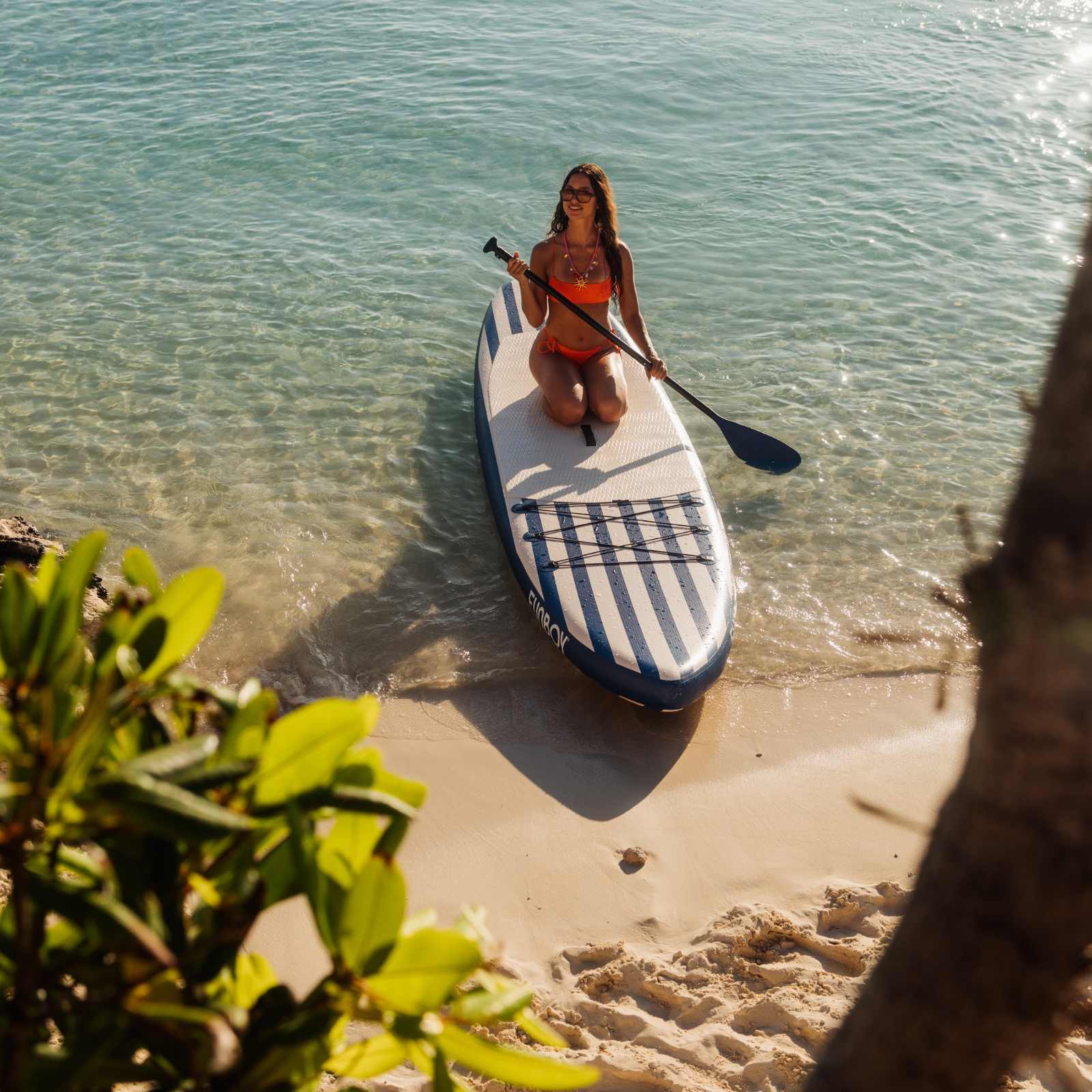 Woman on a striped paddleboard in clear water near a sandy beach.