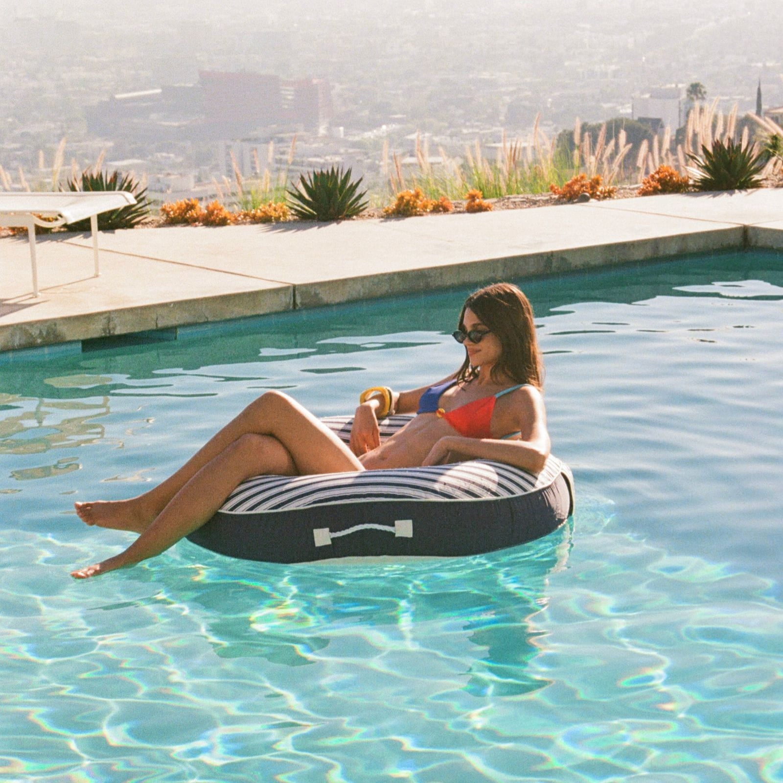 Woman lounging on a striped inflatable tube in a pool with a scenic background