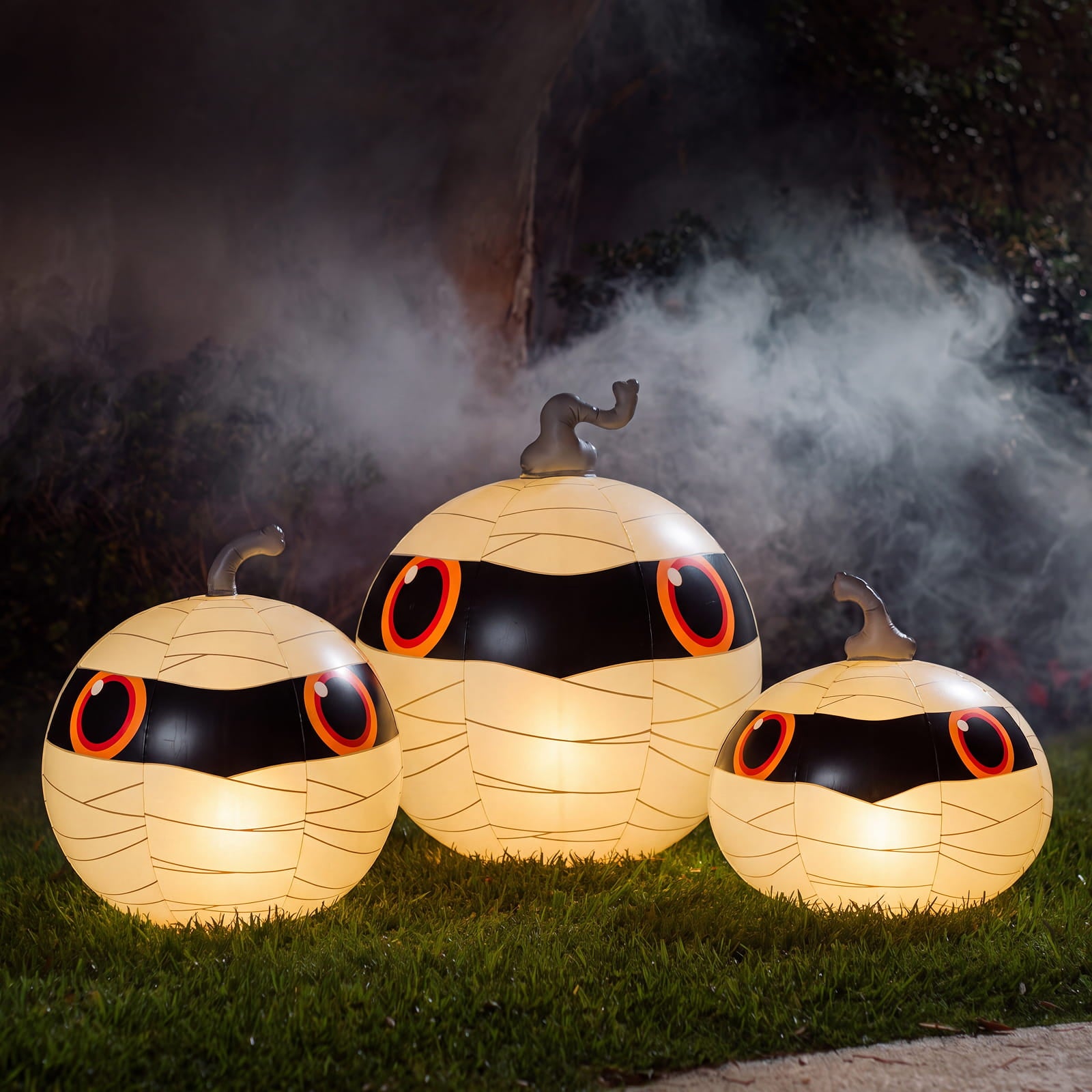 Three glowing mummy-shaped pumpkin decorations with large red and orange eyes sit on grass outdoors, with mist and a dark background.