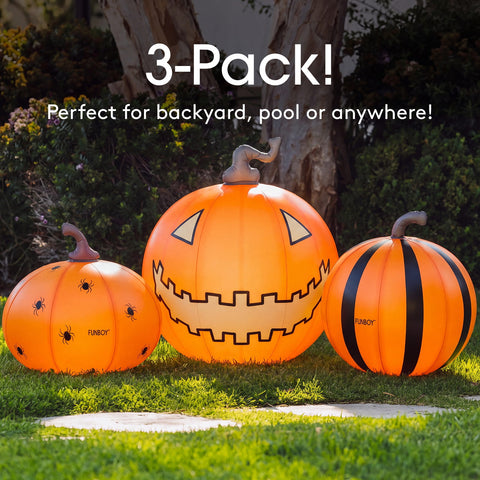 Three orange inflatable Halloween pumpkins displayed outdoors on grass in front of trees. The center pumpkin has a jagged carved mouth and triangular eyes, the left pumpkin has spider holes and the word 'FABCY', and the right pumpkin has black vertical stripes.