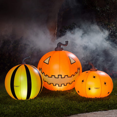 Three inflatable Halloween pumpkins on grass at night with smoke in the background. The left pumpkin is yellow and black striped like a bumblebee, the center pumpkin is bright orange with a jack-o'-lantern face, and the right pumpkin is orange with spiderweb graphics and black spots.