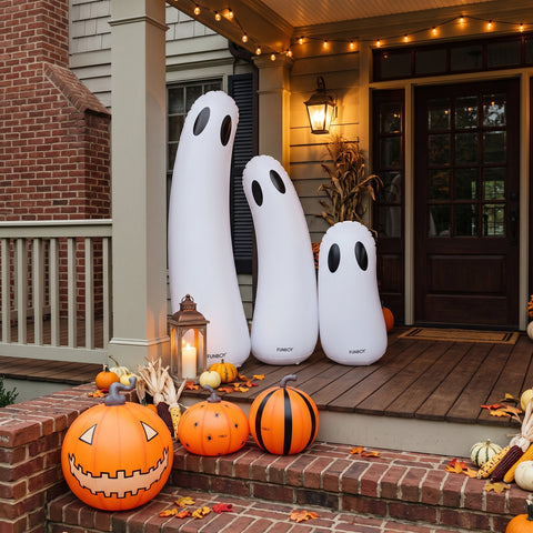 Three white inflatable ghost Halloween decorations of different sizes standing on a wooden front porch, surrounded by pumpkins, fall leaves, and a lantern.
