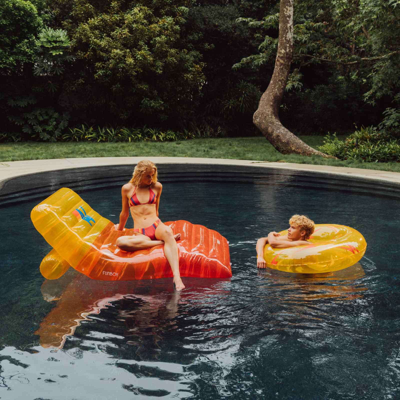 Two people lounging on inflatable rafts in a pool surrounded by greenery. Aviator Nation x FUNBOY