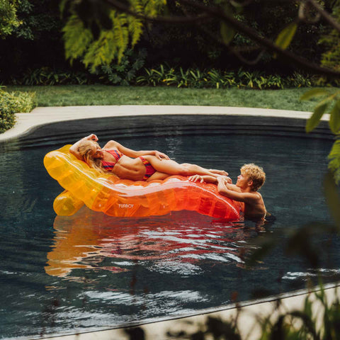 Two people lounging on a colorful inflatable raft in a pool surrounded by greenery. Aviator Nation x FUNBOY