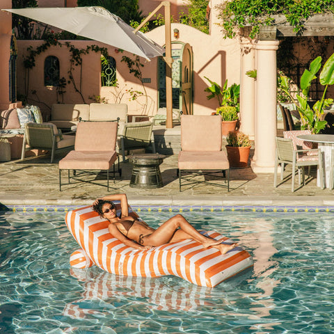 Woman lounging on a striped pool float in a pool area with chairs and umbrellas.