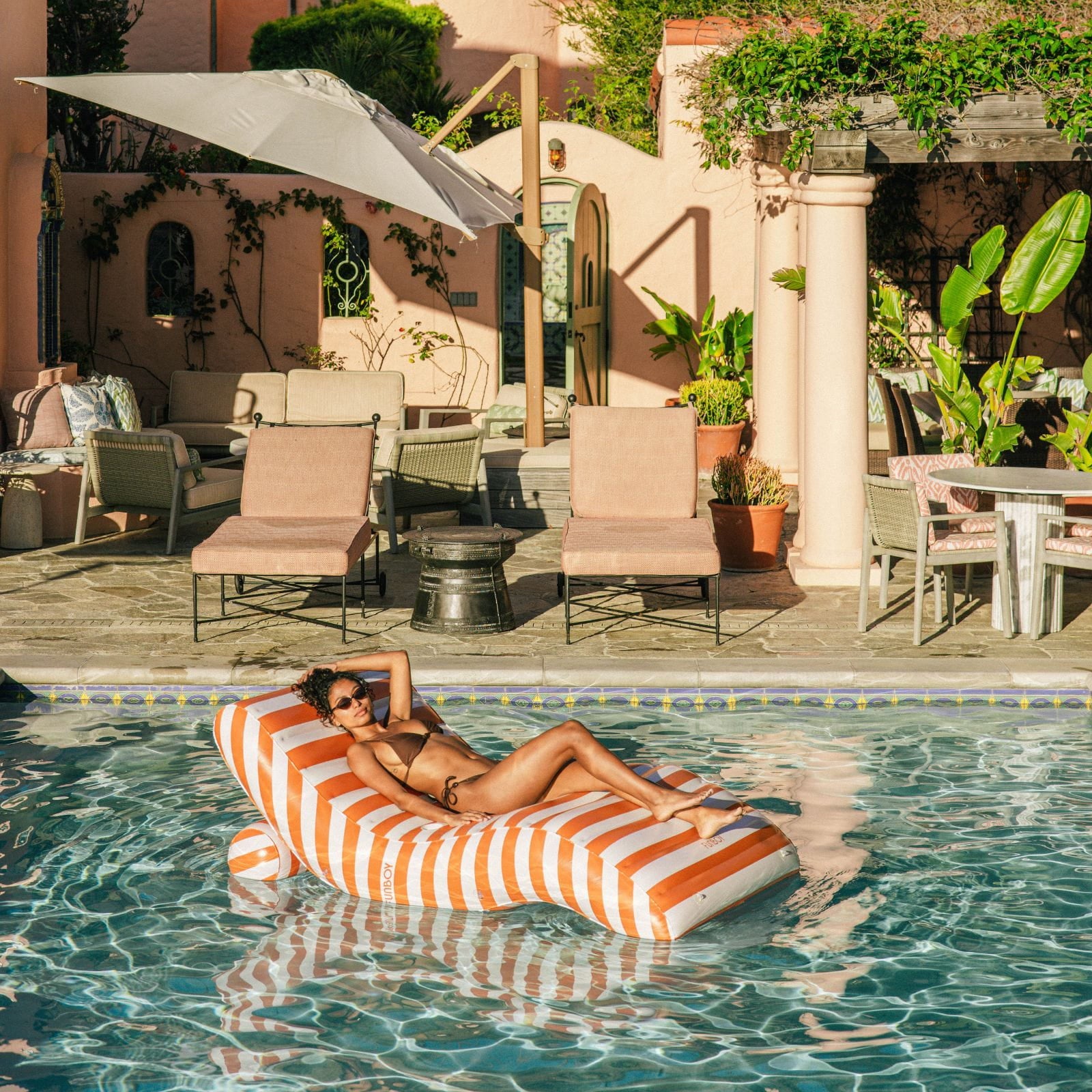 Woman lounging on a striped pool float in a pool area with chairs and umbrellas.