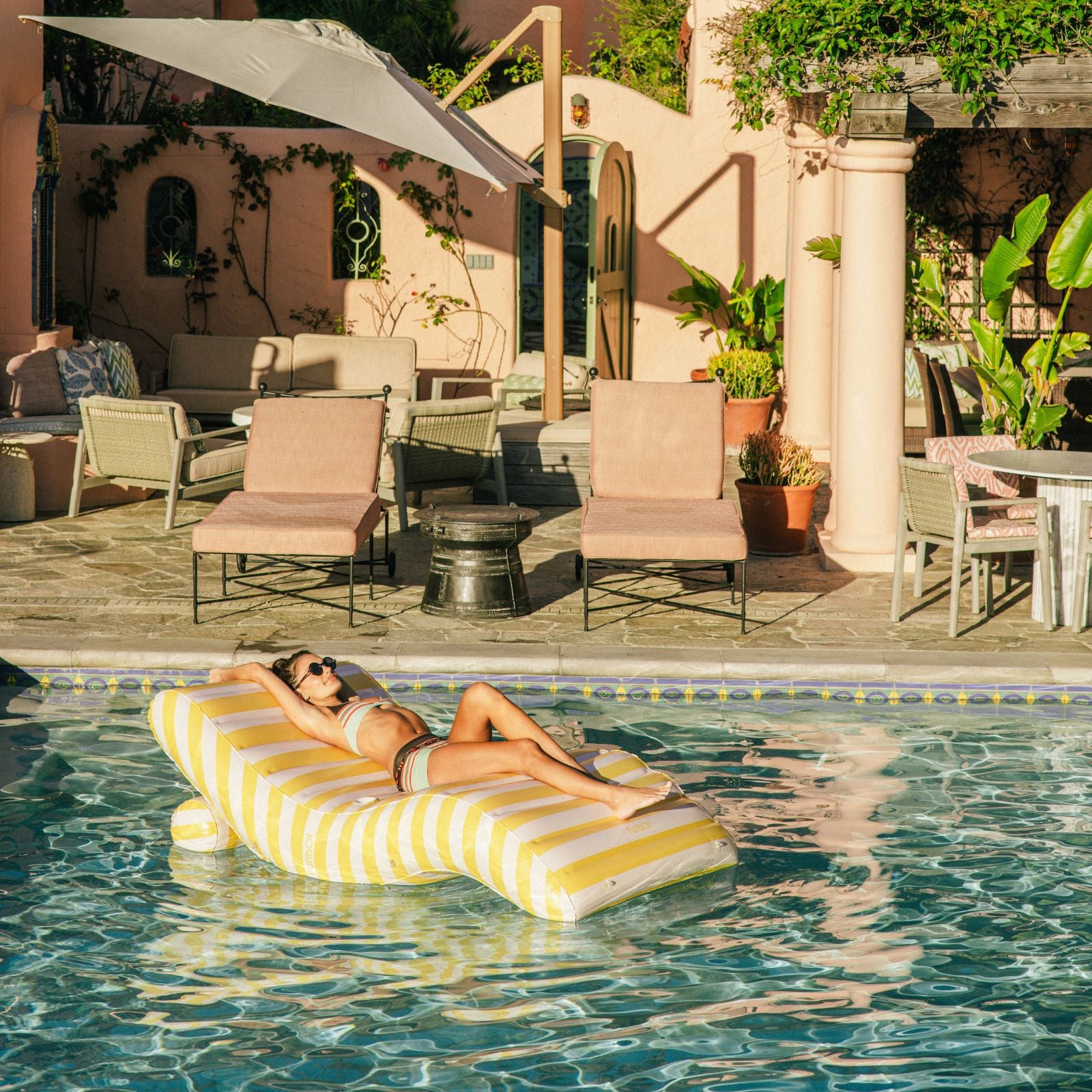Woman lounging on a striped pool float in a pool area with chairs and plants.