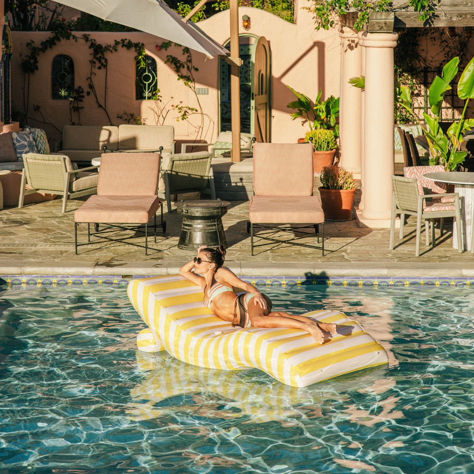 Woman lounging on a yellow and white striped pool float in a pool area with chairs and plants.
