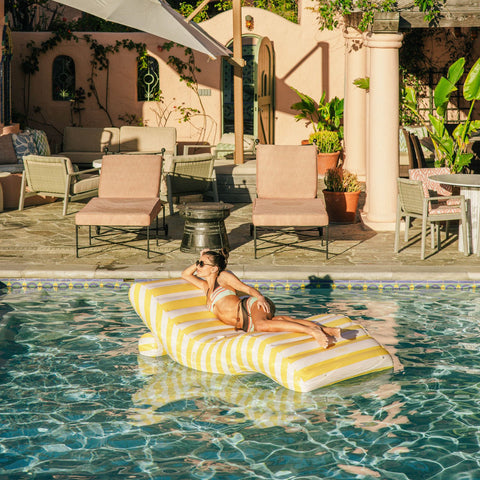 Woman lounging on a yellow and white striped pool float in a pool area with chairs and plants.