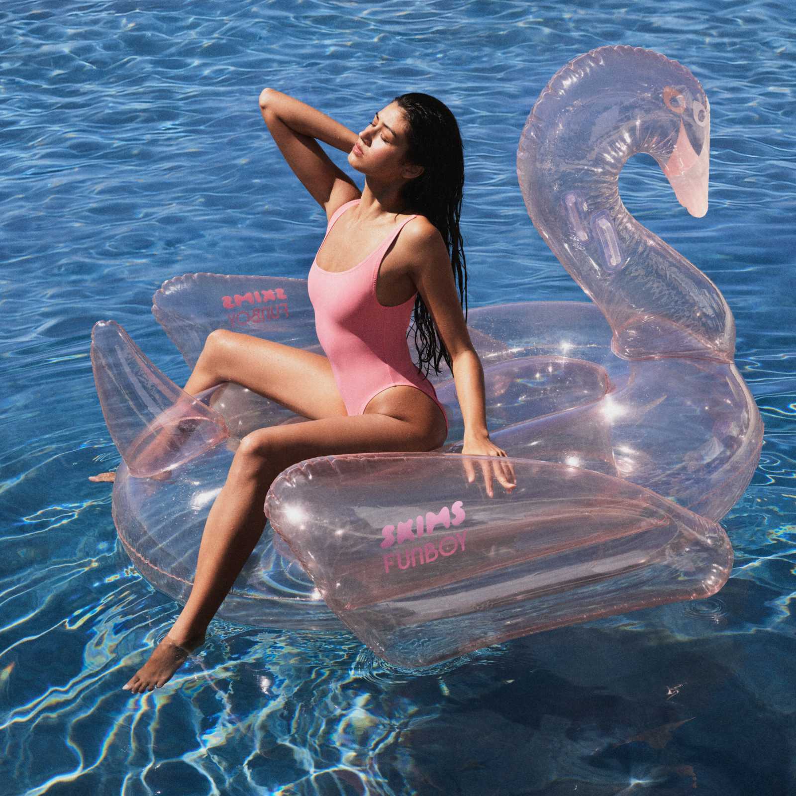 Woman in a pink swimsuit sitting on a clear inflatable swan SKIMS x FUNBOY pool float in blue water.