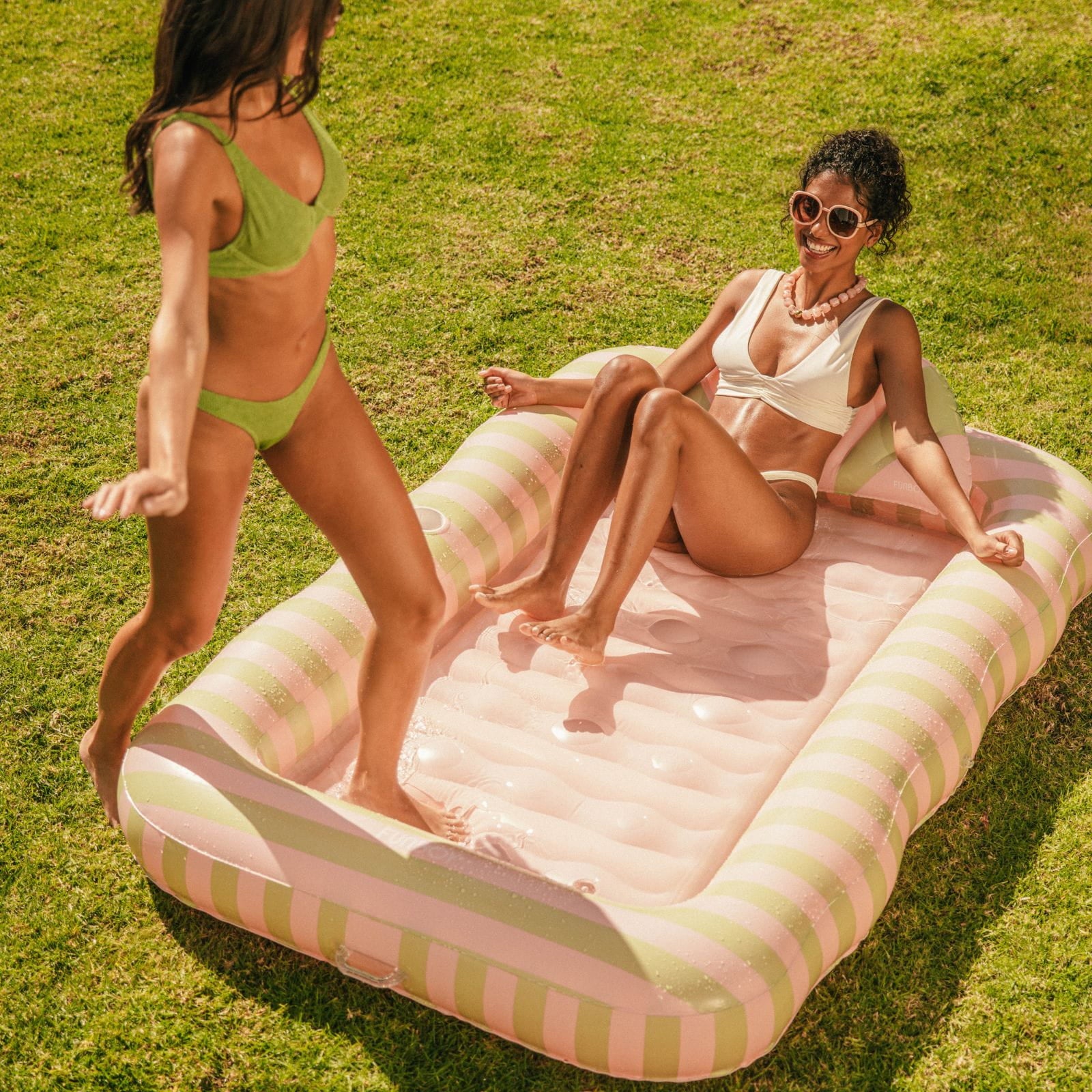 Two women in swimsuits on an inflatable striped pool float in a grassy area