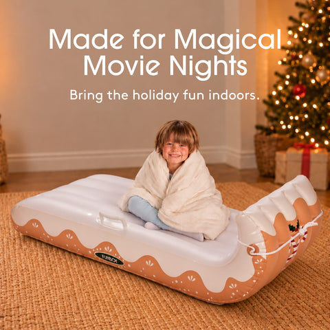 A child wrapped in a white blanket sits on an inflatable holiday-themed bed. The bed is white with peach-colored wavy borders and a gingerbread man design on the headrest.