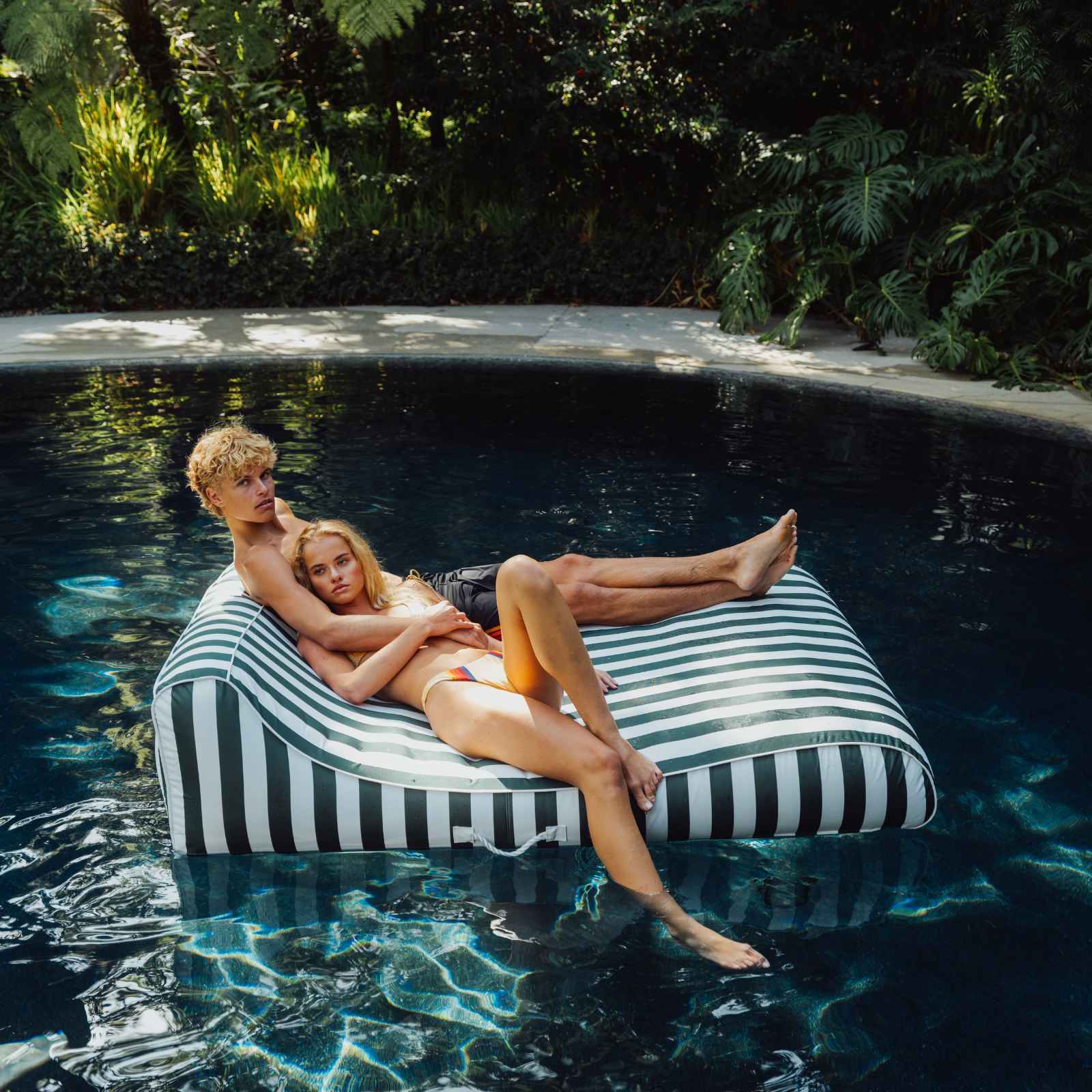 Two people relaxing on a striped inflatable raft in a pool surrounded by greenery. XL Green Fabric Lounger
