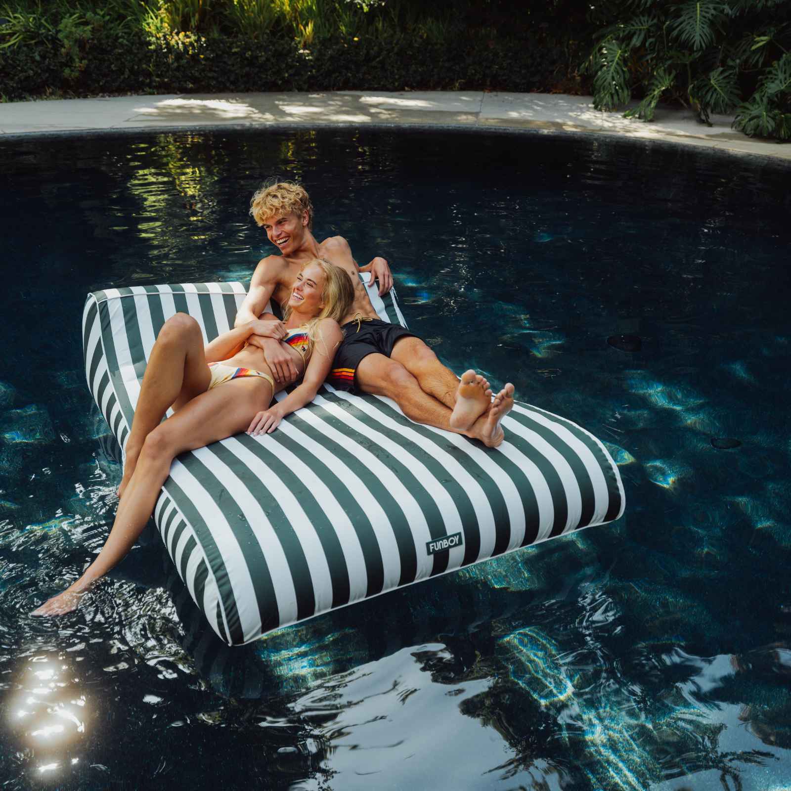 Two people relaxing on a striped pool float in a pool. XL Green Fabric Lounger