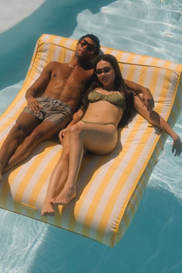 Two people lying on a yellow and white striped pool float in a pool