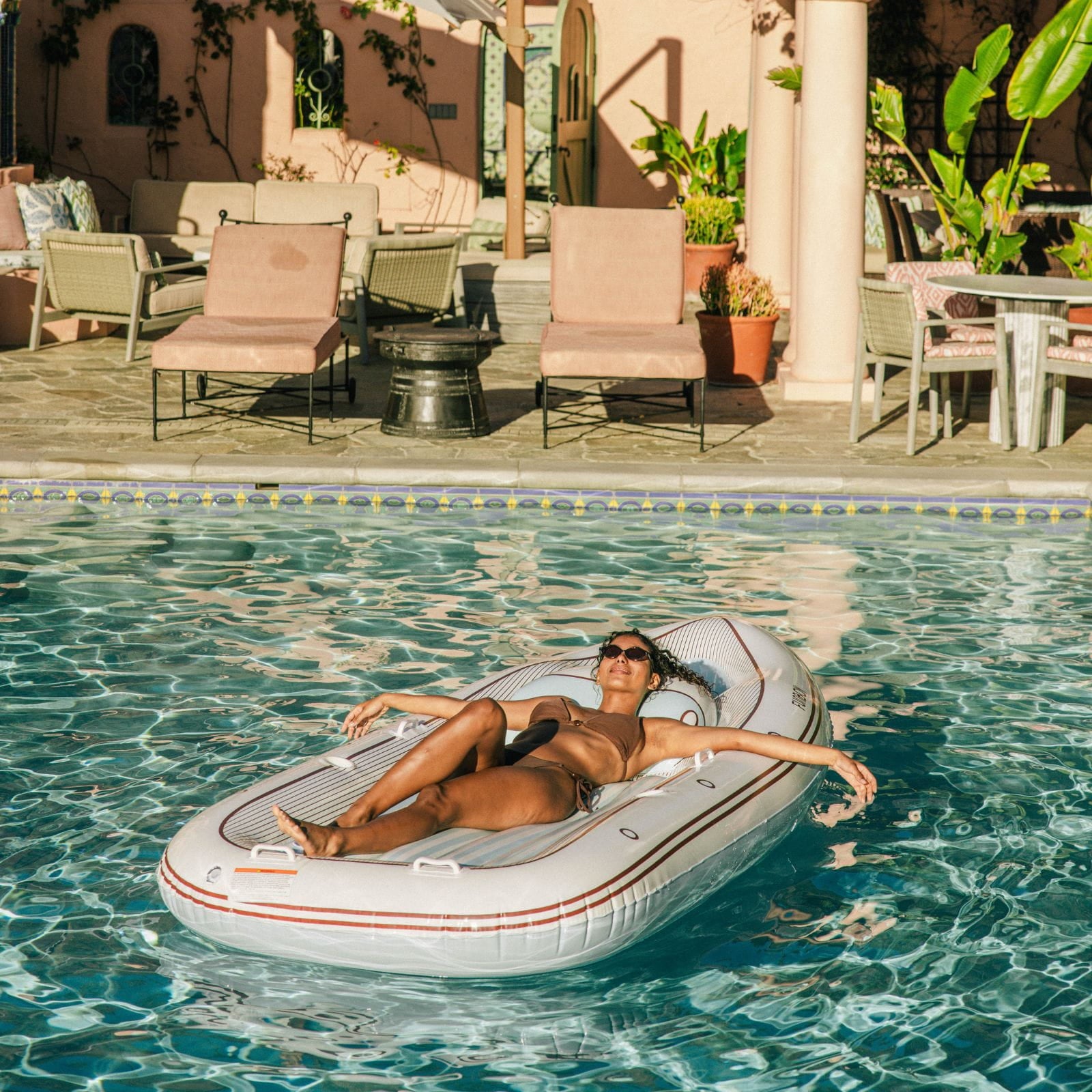 Woman relaxing on an inflatable yacht pool float in a pool with a sunlit patio in the background