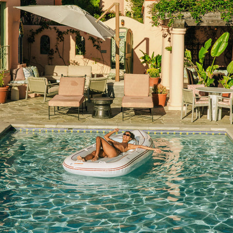Woman relaxing on a pool float in a swimming pool with a patio and outdoor furniture in the background.