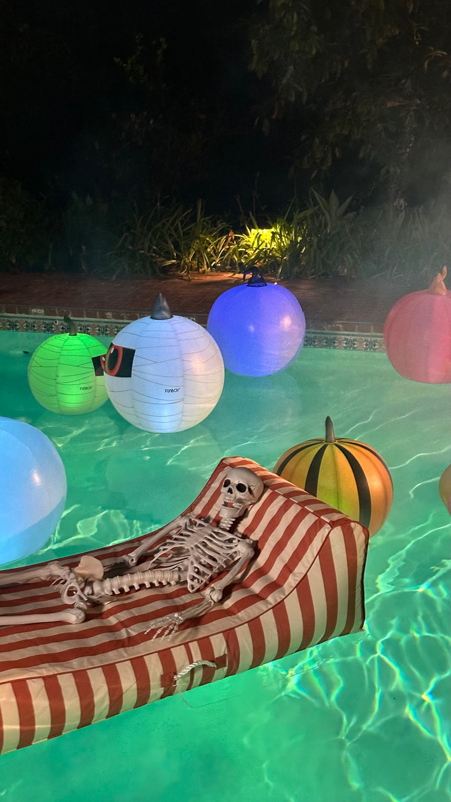 An inflatable skeleton lounge chair floats in a lit swimming pool at night, surrounded by glowing paper lanterns and a striped red-and-white float.