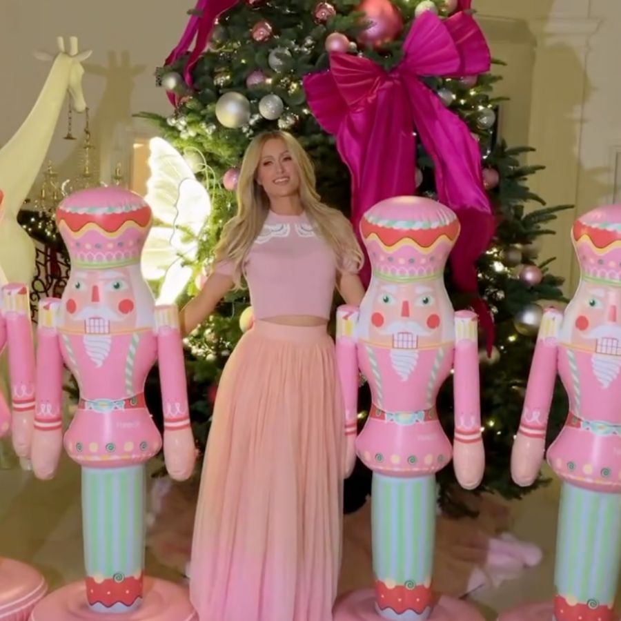 Paris Hilton between two large pink inflatable nutcracker figures in front of a decorated Christmas tree with a big magenta bow.