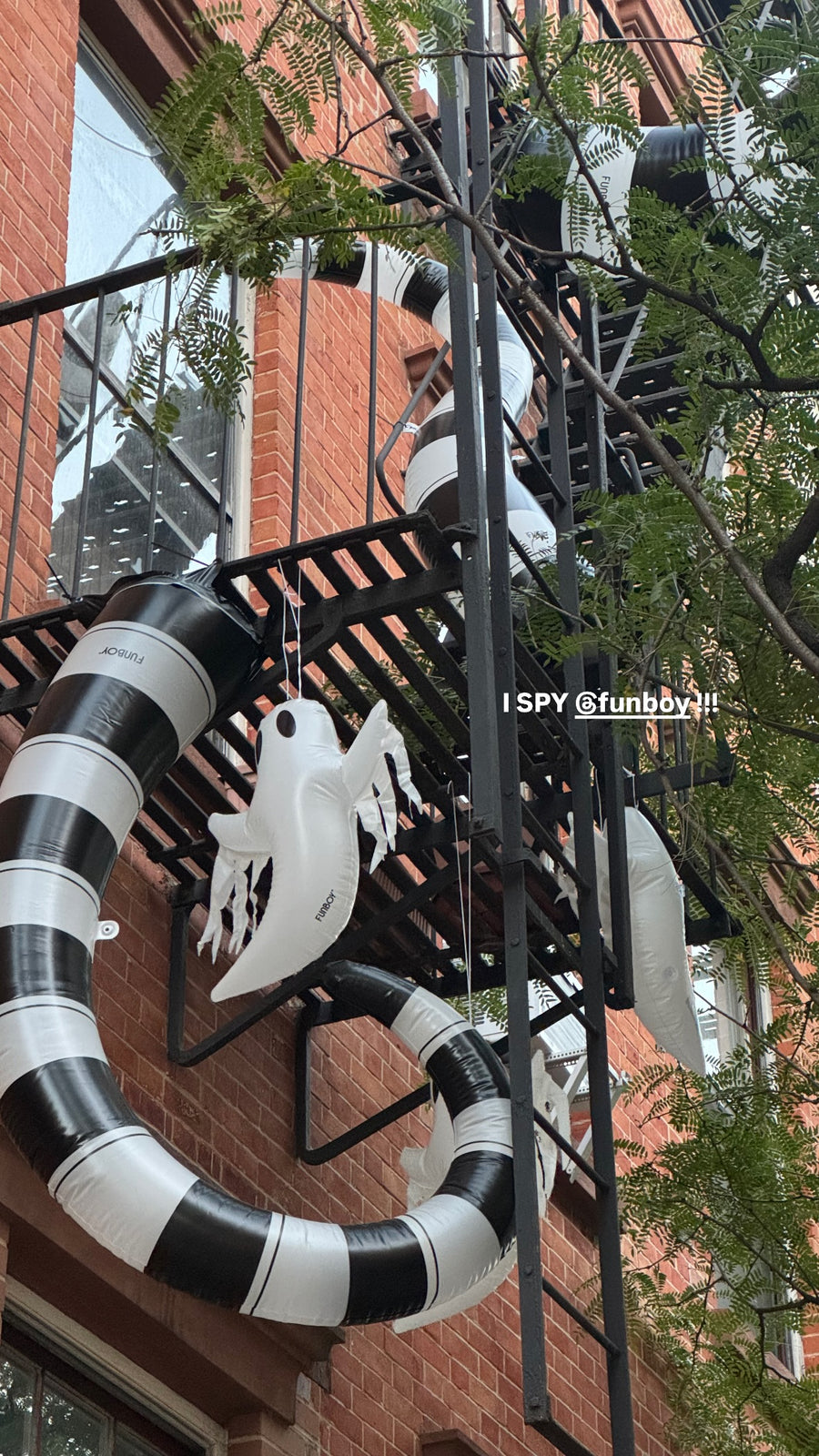 A large black and white striped inflatable snake wraps around a brick building exterior, with a white ghost decoration hanging from the fire escape. Green tree branches overlay the scene.